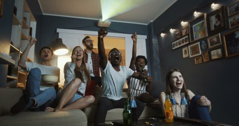 Excited young multiethnic friends get emotional and shout watching sports on TV together at home slow motion. Cheerful students get crazy, raise hands cheering for national team during world cup.